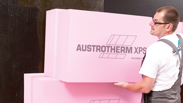 Arbeiter hält rosa Wärmedämmplatten mit Markenaufdruck an Wand.